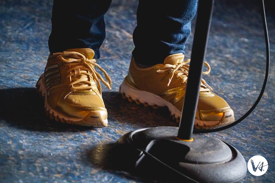 [ID: Gold running shoes and jeans of a person standing at behind a mic and mic stand on stage. /endID]