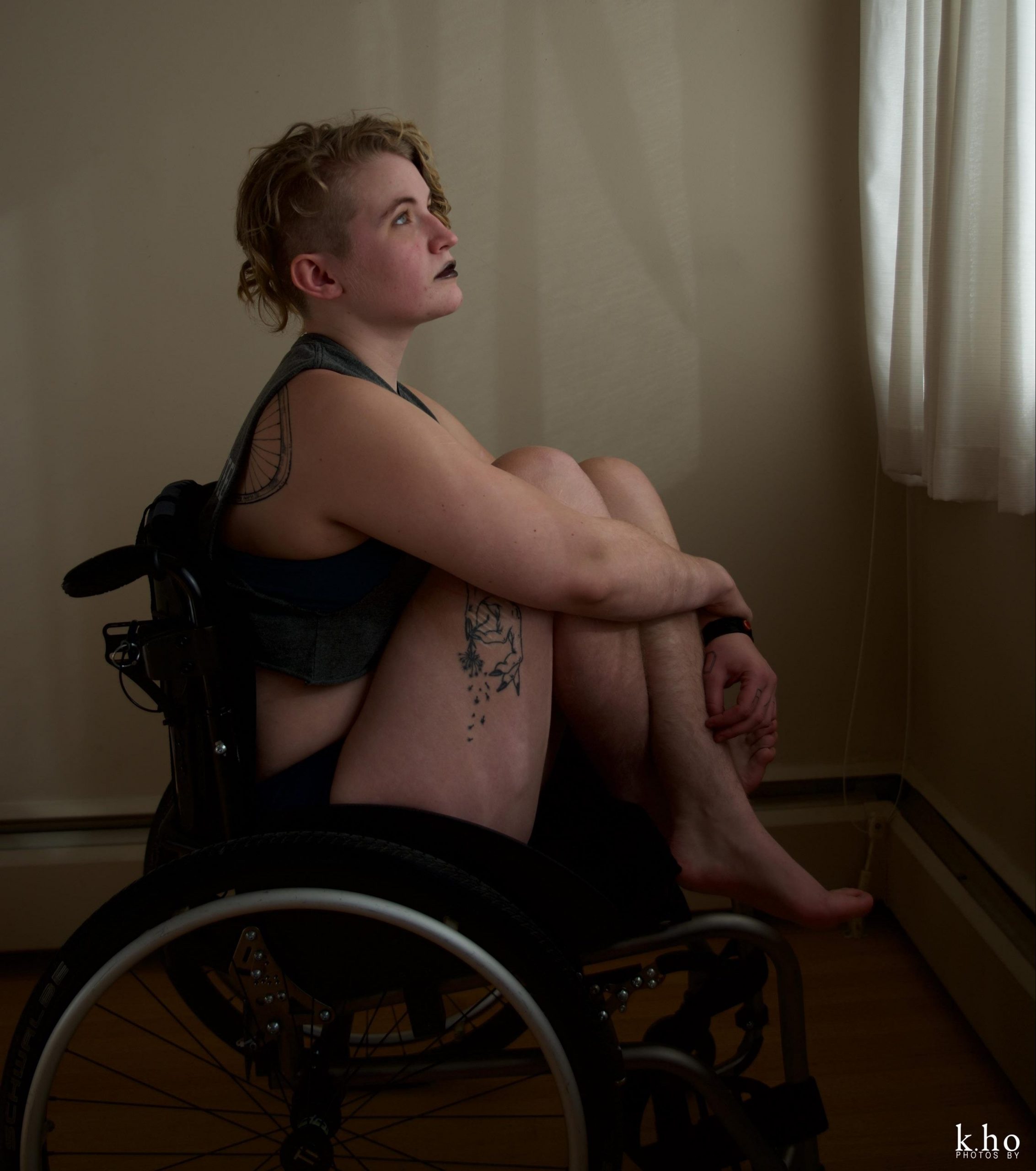 [ID: Q in black lipstick, a grey crop top, and underwear seated in their manual wheelchair, holding their knees to their chest as they look out a window; a bicycle wheel tattoo is partially visible on their shoulder blade, and a hand of fate holding a dandelion puff as the seeds disperse tattoo is on their thigh /end ID]