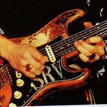 [ID: A close-up of a well-worn wooden electric guitar with SRV initials on it being played by a person's hands with large gold circular rings on the ring fingers. Tha person a black embroidered with white shirt. /end ID]