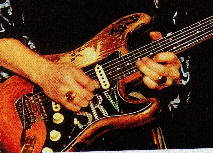 [ID: A close-up of a well-worn wooden electric guitar with SRV initials on it being played by a person's hands with large gold circular rings on the ring fingers. Tha person a black embroidered with white shirt. /end ID]