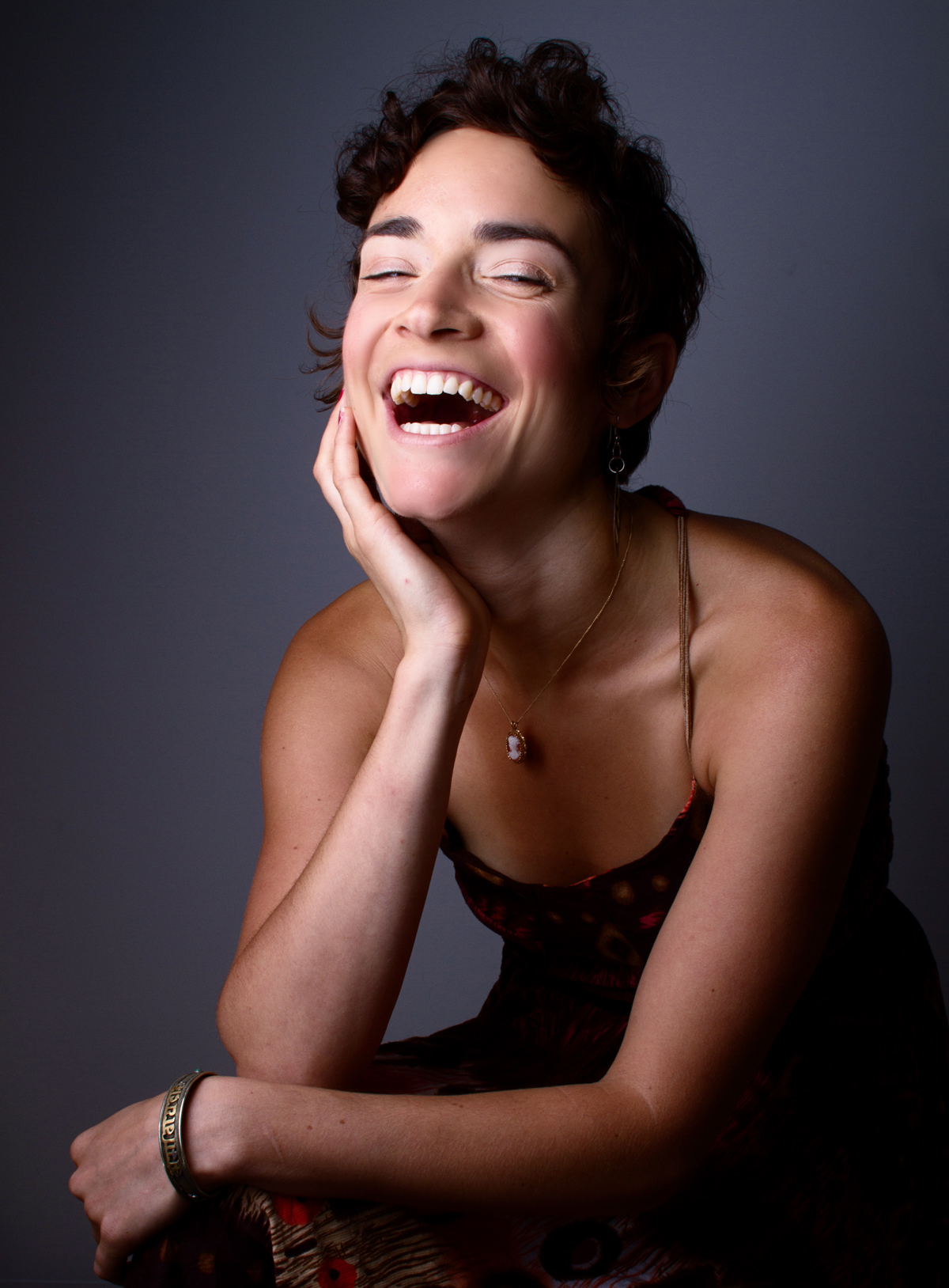 [ID: Moe Clark sits laughing into the camera. She is wearing a camisole style top and a cameo necklace/end ID]