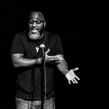 [ID: Christopher Michael is standing on stage speaking into a microphone on a stand in front of him. He is bearded. His eyes are closed behind glasses and his eyebrows are raised. His left arm is stretched toward audience palm up at a 45 degree angle with his right hand indicating the inside of his left elbow. He is wearing a black t-shirt with a black vest over it and jeans. /endID]