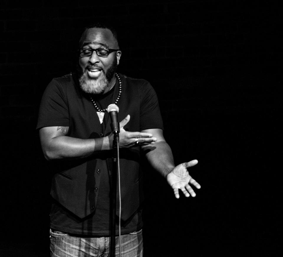 [ID: Christopher Michael is standing on stage speaking into a microphone on a stand in front of him. He is bearded. His eyes are closed behind glasses and his eyebrows are raised. His left arm is stretched toward audience palm up at a 45 degree angle with his right hand indicating the inside of his left elbow. He is wearing a black t-shirt with a black vest over it and jeans. /endID]
