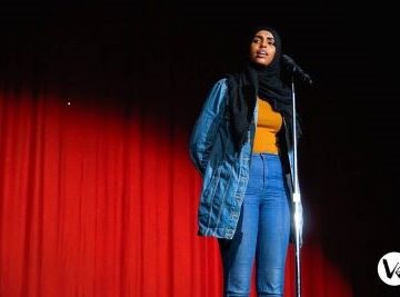 [ID: Ifrah Hussein, 2018 Canadian Individual Poetry Slam champion stands at a microphone on a stand in front of a red curtain illuminated by a spotlight. Her mouth is open speaking. She is looking to her right. She is wearing blue jeans and a denim jacket with a yellow t-shirt under it. She wears a black hijab. Her hands are clasped behind her back. /endID]