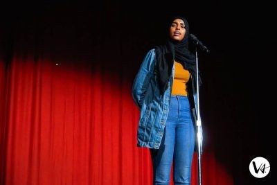 [ID: Ifrah Hussein, 2018 Canadian Individual Poetry Slam champion stands at a microphone on a stand in front of a red curtain illuminated by a spotlight. Her mouth is open speaking. She is looking to her right. She is wearing blue jeans and a denim jacket with a yellow t-shirt under it. She wears a black hijab. Her hands are clasped behind her back. /endID]