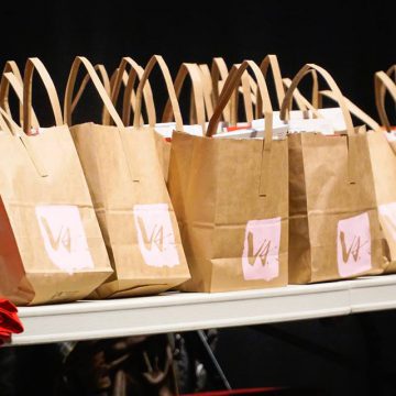 [ID: A row of paper bags with Verses logo screenprinted in pink are lined up along a table. The corner of a stack of red tshirts is on the table at the left bottom edge of the photo. /endID]