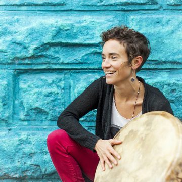 [ID: Moe Clark is crouched in front of a stone wall painted turquoise. Her right hand is on a large hand drum. She is smiling, her face turned to the right. She is wearing a dark grey cardigan and red pants. /endID]