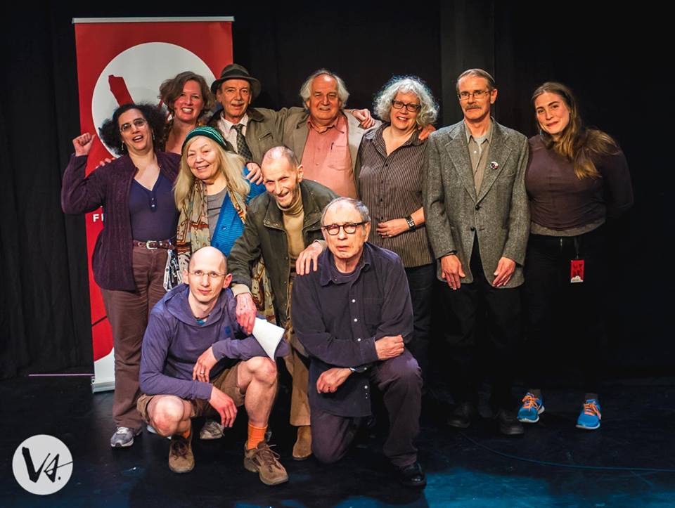 [ID: Group photo of 11 members of Thursdays Writing Collective on stage together. They are all smiling. Two members are crouched at the front of the group for the photo. A red Verses banner is in the background. /endID]