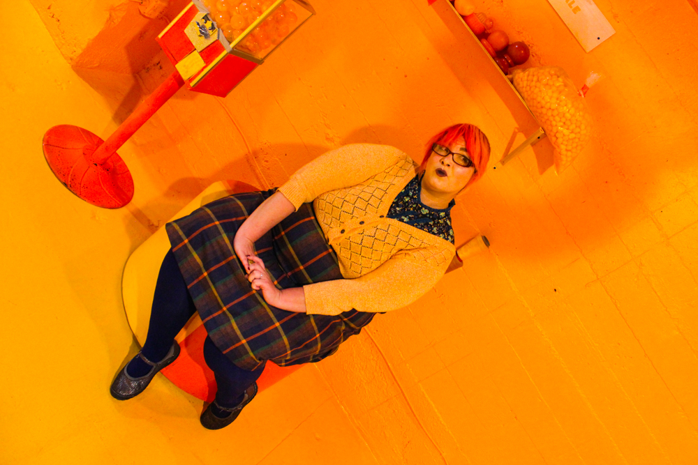 [ID: Kitty Stryker sits on a round orange and yellow ottoman chair. Her feet are tucked to one side. The image is at a tilt and is washed in yellow. Kitty wears a dark plaid skirt, black stockings, Mary-Jane shoes, and a yellow v-necked button up cardigan over a flowered shirt. Her hair is orange. She wears glasses. Her mouth is pursed. Her head is tilted and her eyes are looking to one side. To her right there is an orange gumball machine. Above her on a shelf on the yellow wall behind her, there is a bag of gumballs. /endID]
