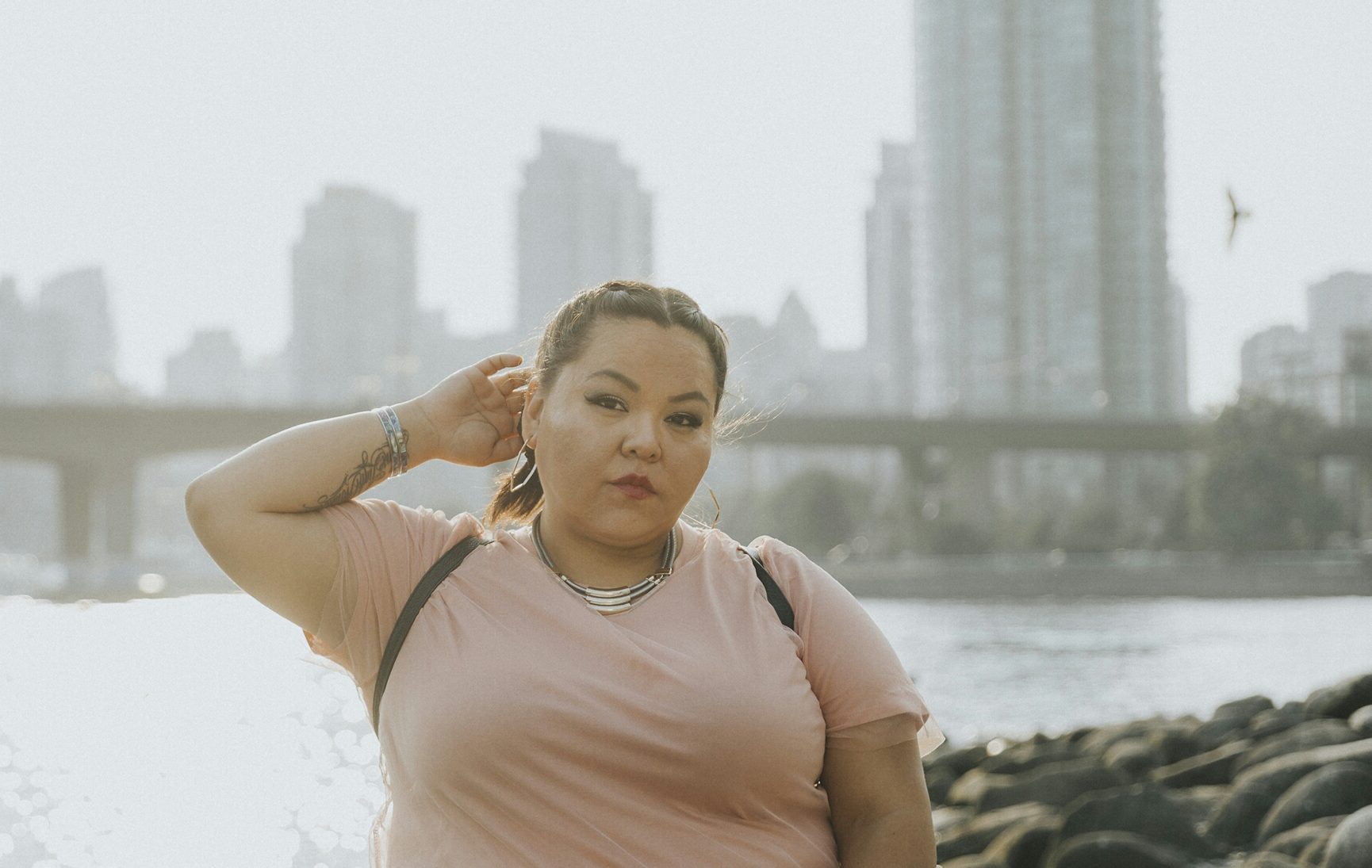 [ID: JB the First Lady stands with the seashore, water, a bridge, and Vancouver skyline behind her. She wears a light pink shirt. Her right hand is touching the back of her head behind her right ear. Her hair is braided. /endID]